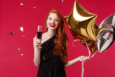 Celebration, holidays and party concept. Carefree gorgeous redhead woman having fun with friends throwing birthday, wear black dress, hold balloons and drink champagne with confetti in airの写真素材