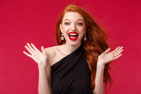 Makeup, beauty and women concept. Close-up portrait of gorgeous redhead woman happy to see someone at her party of event, clap hands surprised and pleased, standing red backgroundの写真素材