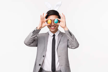 Holiday, people and celebration concept. Waist-up portrait of funny, playful and enthusiastic asian guy in grey suit and tie, holding painted eggs over eyes and smiling, enjoy Easter partyの写真素材