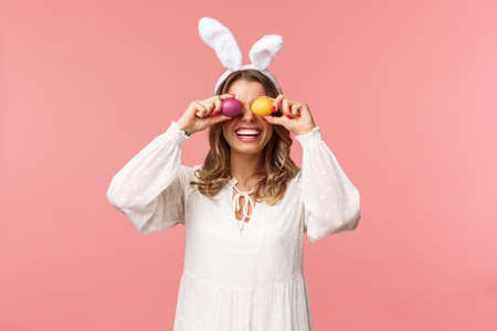 Holidays, spring and party concept. Portrait of lovely, tender smiling woman in rabbit ears and white dress celebrating Easter day, holding painted eggs on eyes and grinning, pink backgroundの写真素材