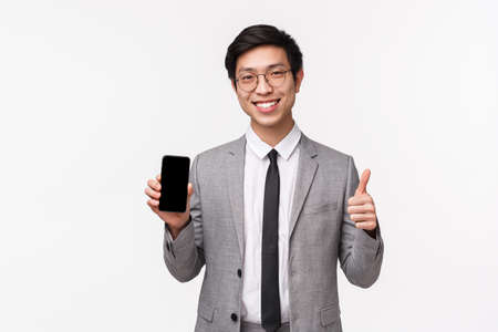 Waist-up portrait of satisfied good-looking asian young man in grey suit, show mobile phone display and thumbs-up, approval cool new application, recommend buy subscription, white backgroundの写真素材