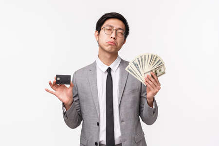 Waist-up portrait of bored and reluctant young rich asian guy in grey suit, feel gloomy and tired of wasting money, holding big sum of cash, dollars and credit card, complaining, white backgroundの写真素材