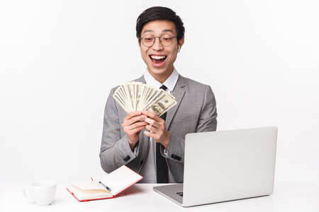 Portrait of successful asian businessman sitting at his office counting money, holding fan of dollars grinning delighted, earn big sum of cash on recent deal, sit near laptop, wear wireless earbudsの写真素材