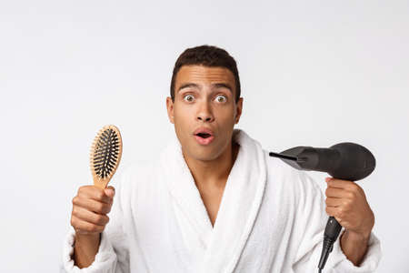 Portrait of mature funny beautiful tan-skinned african guy with curly hair in casual white bathrobe having problems with combing hair and hair dryerの写真素材