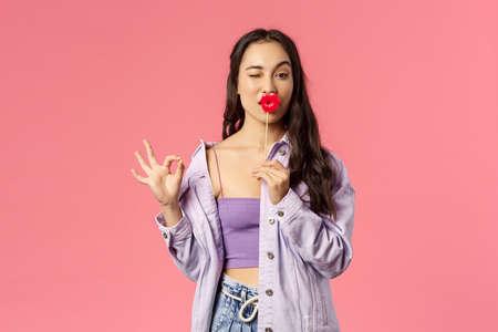 Holidays, lifestyle and people concept. Portrait of funny, sassy mixed-race girl in stylish outfit, show okay no problem sign, hold stick of big lips, wink cute camera, standing pink backgroundの写真素材