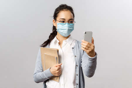 Covid19, virus, health and medicine concept. Young asian girl in medical mask, holding notebooks and work reports, taking selfie on smartphone, videocalling friends while social distancingの写真素材