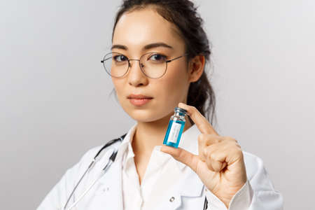 Close-up portrait of serious-looking young asian infectionist, female doctor showing ampule with coronavirus vaccine, invented covid19 medicine, look determined, start treating patientsの写真素材