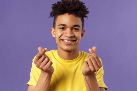 Close-up portrait of lovely cute young man with afro haircut, showing korean heart finger signs and...