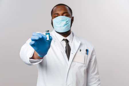 Covid19, pandemic and healthcare concept. Serious african-american doctor in white coat, latex gloves and medical face-mask showing new vaccine from coronavirus outbreak, treat peopleの写真素材