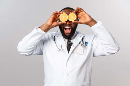 Healthcare, healthy diet and disease concept. Wow so many vitamins. Portrait of amazed happy african-american doctor ask self-distance and eat more vitamins, hold oranges like eyesの写真素材