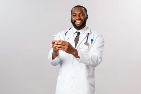 Covid19, pandemic and online medicine concept. Happy friendly african-american doctor using mobile phone and smiling pleased, hold smartphone, receive appointment info in applicationの写真素材
