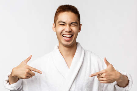 Beauty, spa and leisure concept. Portrait of successful, happy handsome asian man pointing fingers at himself and laughing, talking own accomplishment, wear bathrobe, white backgroundの写真素材