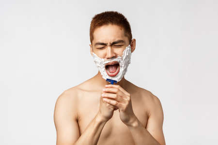 Beauty, people and hygiene concept. Portrait of carefree handsome asian named man shaving and singing, get ready in bathroom, apply cream, sing into razor like microphone, stand white backgroundの写真素材