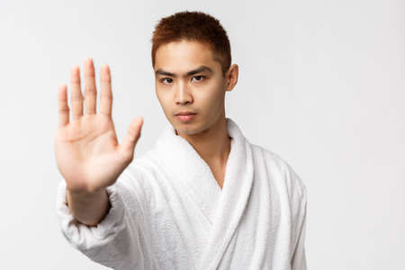 Beauty, spa and leisure concept. Portrait of serious-looking asian man in bathrobe pull hand forward to say stop, tell no to something bad, stay at home during covid19 quarantine, prohibit actionの写真素材