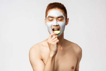 Beauty, skincare and spa concept. Portrait of tender and satisfied asian naked man close eyes, eating cucumber with delight and temptation, wear face mask to treat pimples and blemishesの写真素材