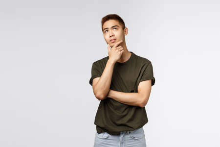 Portrait of young asian man thinking and looking up with thoughtful, serious expression, rub chin pondering choice, need make difficult decision, standing puzzled grey backgroundの写真素材