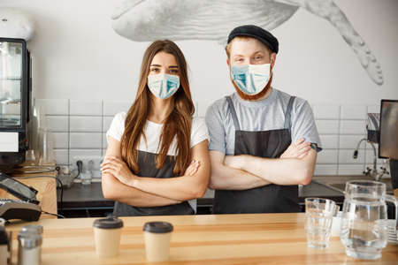 Coffee Business Concept - Barista couple in apron and face mask looking at camera ready to give Coffee Service at the modern coffee shopの写真素材