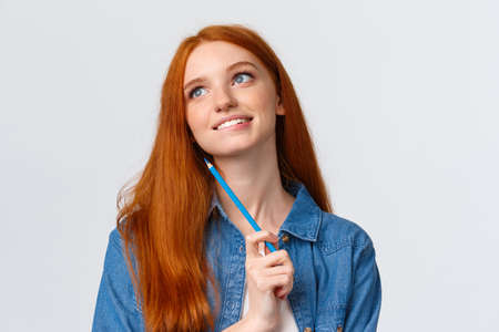 Lovely and creative, thoughtful dreamy smiling redhead woman looking up and thinking, imaging something beautiful, holding colored pencil, drawing, attend art courses, white backgroundの写真素材