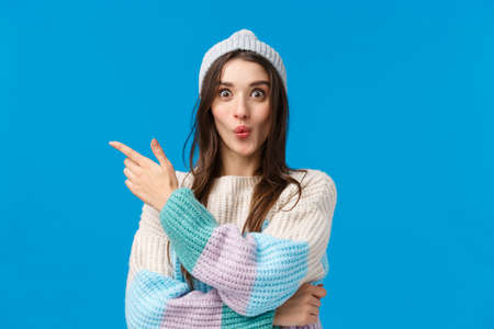 Wow its interesting, lets see. Intrigued and excited cute smiling woman in winter sweater, hat, folding lips curiously pointing finger left, telling about awesome event, standing blue backgroundの写真素材