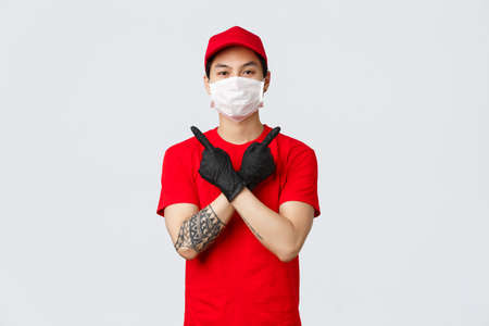 Young confident delivery guy in red t-shirt and cap, wear protective gloves and medical mask to deliver parcels at customer home during covid-19, showing variants, pointing sideways, two choicesの写真素材