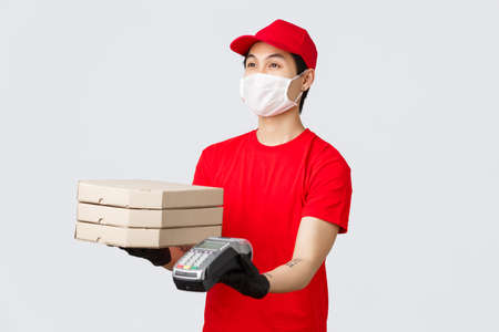 Contactless delivery, safe purchase and shopping during coronavirus concept. Friendly courier in red uniform cap and t-shirt, giving client pizza delivery order and payment terminal, grey backgroundの写真素材