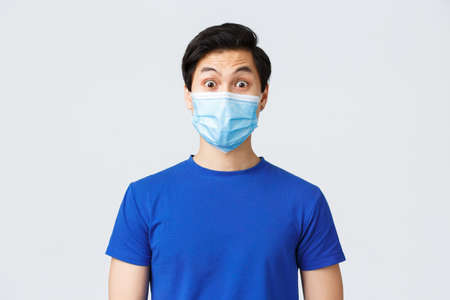 Different emotions, lifestyle and leisure during coronavirus, covid-19 concept. Surprised expression of asian guy in medical mask. Close-up guy raise eyebrows in surprise, listen newsの写真素材