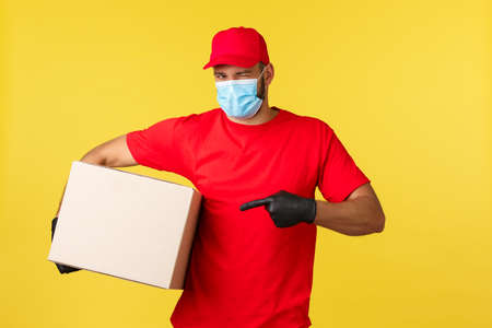 Express delivery during pandemic, covid-19, safe shipping, shopping concept. Cheeky handsome courier bring your parcel, wink and pointing box, wear medical mask protecting from coronavirus spreadの写真素材