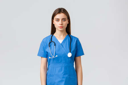 Healthcare workers, prevent virus, insurance and medicine concept. Serious-looking young nurse, doctor in blue scrubs and stethoscope, looking at camera, standing grey backgroundの写真素材