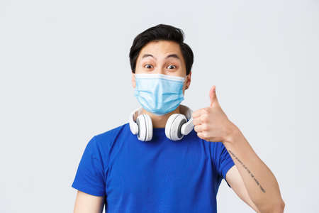 Different emotions, lifestyle and leisure during coronavirus, covid-19 concept. Close-up of upbeat asian guy in medical mask show thumb-up in approval of awesome new song playing in headphonesの写真素材