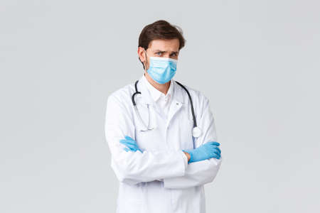 Hospital, healthcare workers, covid-19 treatment concept. Miserable and sad young doctor feeling uneasy, wear medical mask, scrubs and gloves, frowning look jealous or offended, stand grey backgroundの写真素材