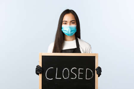 Small business, covid-19 pandemic, preventing virus and employees concept. Friendly asian female employee inform cafe visitors or store clients that we are closed, holding sign, wear medical maskの写真素材