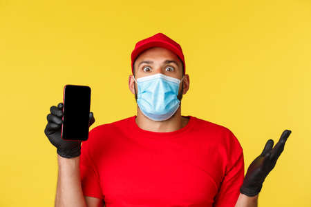 Covid-19, delivery, shopping, contactless payment and social distancing concept. Close-up of astonished, excited courier talking about new application, show mobile phone screen, wear medical maskの写真素材