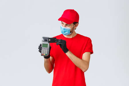 Contactless delivery, payment and online shopping during covid-19, self-quarantine. Courier in red uniform and gloves with face mask, showing POS paying terminal and credit card, pay for orderの写真素材