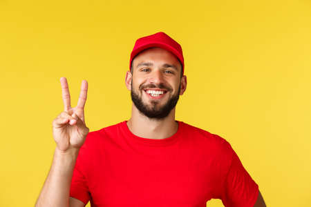 Close-up friendly cheerful, bearded delivery guy show peace sign, wear red uniform cap and t-shirt, courier likes taking care customer orders, safely shopping and parcel transfer, yellow backgroundの写真素材