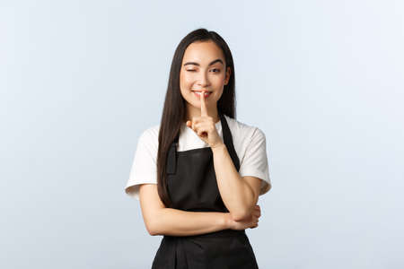 Coffee shop, small business and startup concept. Cheeky young asian female barista prepared surprise for cafe clients, wink and make hush gesture with finger pressed to lips, smilingの写真素材