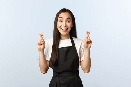 Coffee shop, small business and startup concept. Optimistic cute asian waitress cross fingers good luck. Upbeat barista in black apron making wish, praying or pleadingの写真素材