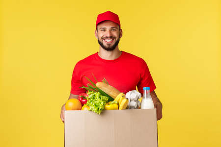 Online delivery, shopping and food order concept. Nice delivery guy, courier in red uniform company, bring grocery order in box to client, smilign friendly, standing yellow backgroundの写真素材