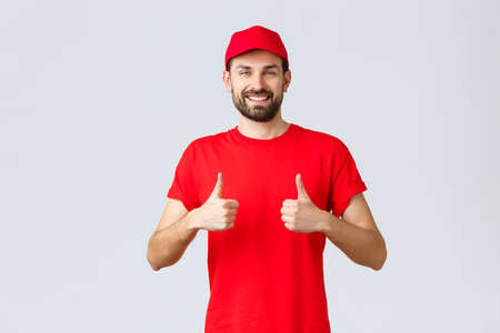 Online shopping, delivery during quarantine and takeaway concept. Cheerful courier in red uniform cap and t-shirt, recommends make orders, thumbs-up in approval, grey backgroundの写真素材