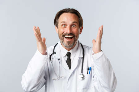 Covid-19, preventing virus, healthcare workers and vaccination concept. Close-up of excited bearded male doctor in white coat raising hands up thrilled, hear great news and rejoicingの写真素材