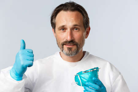 Covid-19, preventing virus, healthcare workers and vaccination concept. Middle-aged male doctor in personal protective equipment, take-off respirator and show thumb-up, assure all under controlの写真素材