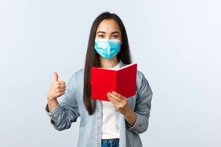 Covid-19 pandemic, education during coronavirus, back to school concept. Pleased happy asian girl in medical mask show thumb-up as checking friend homework task in notebookの写真素材