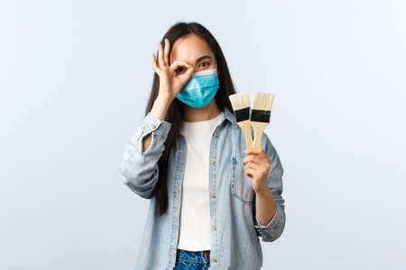 Social distancing lifestyle, covid-19 pandemic, self-isolation hobbies and leisure concept. Cute pretty asian girl in medical mask show okay sign over eye and painting brushesの写真素材