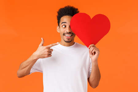 Excited happy, charismatic african-american male in white t-shirt, holding heart valentines card over face, pointing at sign and smiling amused, prepared surprise gift for lover, orange backgroundの写真素材