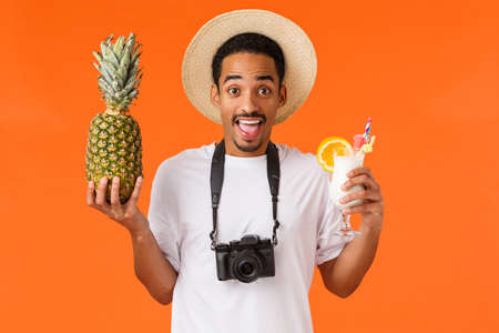 Cheerful happy young man partying enjoying travelling hot country, paradise island, booked tickets travel agency, holding pineapple and cocktail, taking pictures on camera, orange backgroundの写真素材