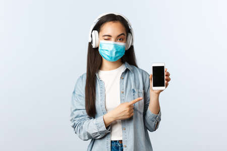 Social distancing lifestyle, covid-19 pandemic and self-isolation leisure concept. Cheeky modern asian girl in medical mask, listen music wireless headphones, pointing smartphone displayの写真素材