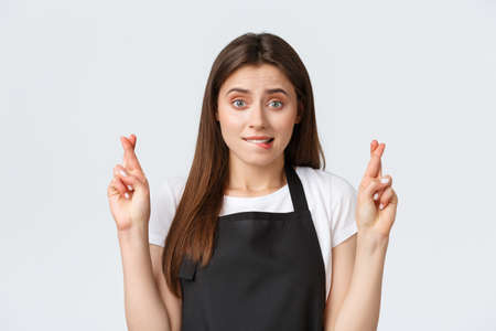 Employees, job employment, small business and coffee shop concept. Nervous cute cafe worker, saleswoman in black apron biting lip worried and cross fingers good luck, making wish, waiting for resultsの写真素材
