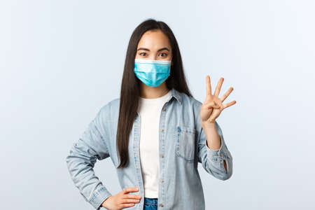 Social distancing lifestyle, covid-19 pandemic and people emotions concept. Smiling carefree asian female in medical mask show four fingers, make order or reservationの写真素材