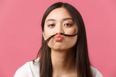 Beauty, people emotions and summer leisure concept. Close-up of funny and bored silly asian girl making fake moustache with hair strand over lip, stare camera playful, pink backgroundの写真素材