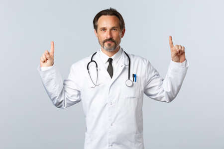 Covid-19, coronavirus outbreak, healthcare workers and pandemic concept. Confident adult doctor in white coat, stethoscope, pointing fingers up and smiling, prescribe pills, advertise productの写真素材