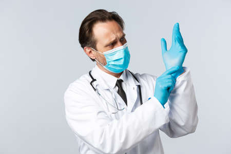 Covid-19, preventing virus, healthcare workers and vaccination concept. Serious professional doctor in medical mask put on gloves before examining patient in clinic, working in hospitalの写真素材